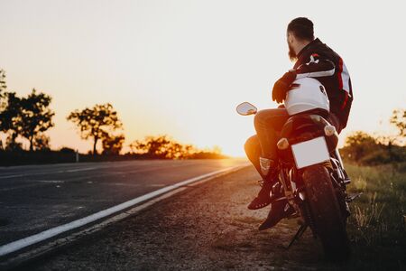 Confident attractive european biker sitting on his bike and looking at the sunset contemplating while resting in his traveling on the bike.の写真素材