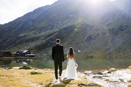 Full length back view of unrecognizable groom and bride sitting on the edge of a lake in the...