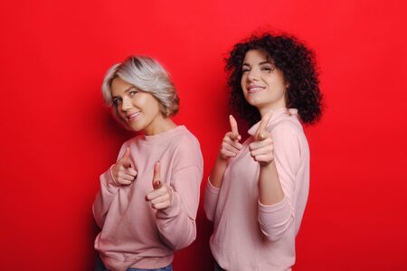 Lovely caucasian lady with curly hair and her blonde friend pointing happily to you on a red backgroundの写真素材