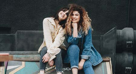 A charming lady with curly hair is posing and smiling near her brunette friend dressed in blue sitting on something outsideの写真素材