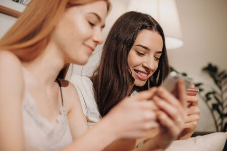Charming caucasian girl with red hair and freckles is texting with her brunette friend sitting on a sofaの写真素材