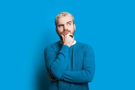Blonde caucasian youngster touching his chin and looking aside while posing on a blue background in...