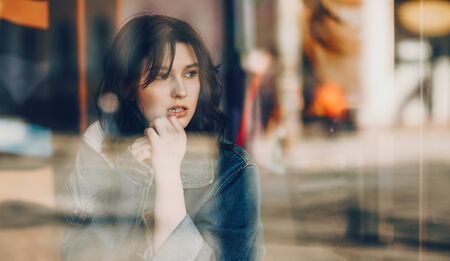 Lovely caucasian girl with black hair and blue jacket is waiting something in a coffeeshop looking away through the windowの写真素材