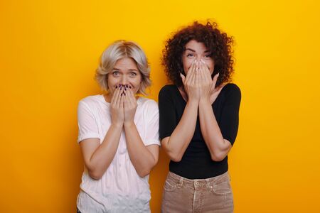 Caucasian curly haired brunette is feeling surprised near her blonde sister covering their mouths...