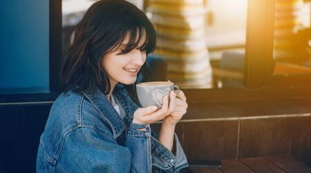 Smiling caucasian girl with short hair and blue jacket is drinking a coffee outside near a coffee shopの写真素材