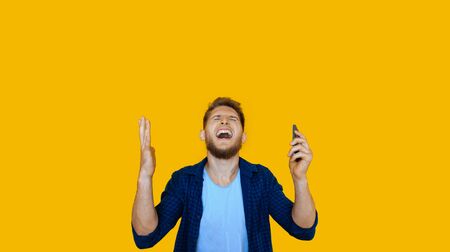 Bearded caucasian man using a phone is gesturing up after winning something on a yellow wall
