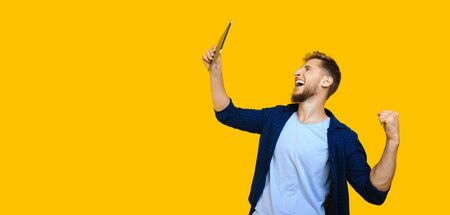 Caucasian man with beard and red hair is looking at his tablet and shouting being happy and posing on a yellow background with freespaceの写真素材