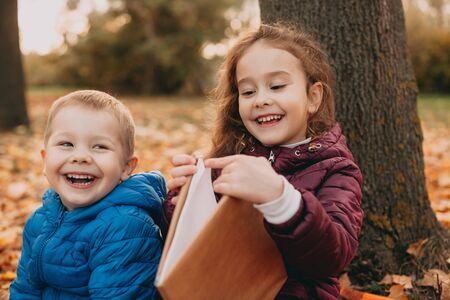 Two caucasian siblings sitting on the ground the park surrounded by leaves are smiling and reading a book togetherの写真素材