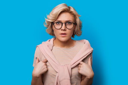 Blonde caucasian lady looking through eyeglasses and pointing to herself while posing on a blue wallの写真素材