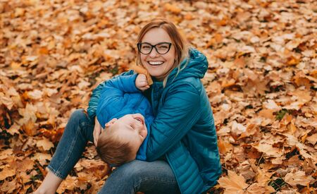 Red haired caucasian mother playing with son while sitting on the fallen colorful leaves in a parkの写真素材