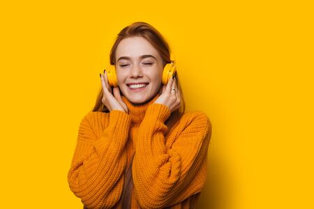 Toothy caucasian girl with red hair and freckles is listening to music with headphones on a yellow...