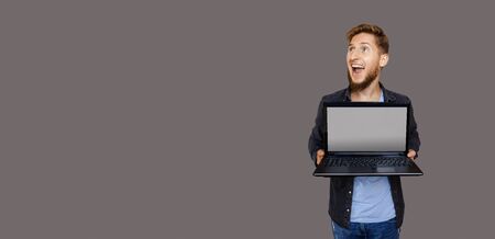 Ginger caucasian man with beard is looking up to gray blank space while holding a laptop and advertising somethingの写真素材