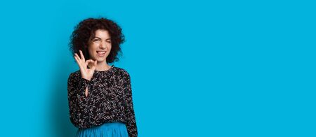 Curly haired caucasian girl is gesturing the okay sign while posing near blue free space