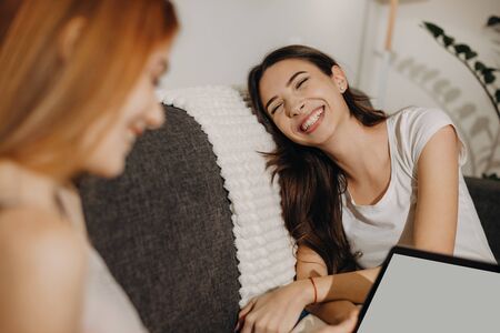 Caucasian ginger girl is using a laptop while having fun with her brunette sister on the sofaの写真素材