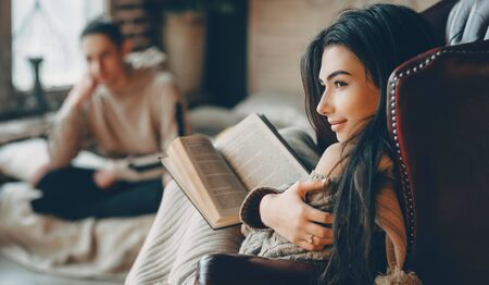 Wonderful caucasian girl with black hair is reading a book while her lover is sitting on the sofa...
