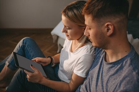 Blonde caucasian woman and her lover are sitting on the floor and using a tablet during a weekend...