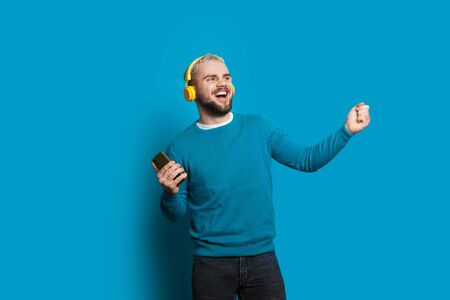 Blonde caucasian man using a phone and headphones is dancing and gesturing on a blue wall