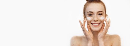 Ginger caucasian lady with freckles is posing on a white wall while applying a special cream on her faceの写真素材