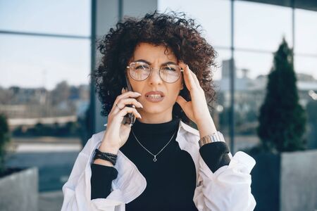Thoughtful caucasian entrepreneur with curly hair and eyeglasses is posing while speaking on phone