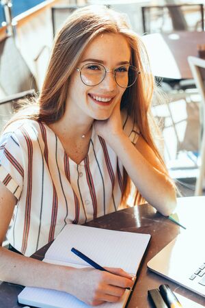 Delightful student with red hair and freckles making some notes while wearing eyeglasses and using laptop in a cafeteriaの写真素材
