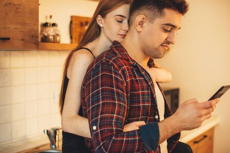 Lovely caucasian couple embracing in the kitchen while he is using his phoneの写真素材