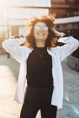 Delight curly haired caucasian woman with eyeglasses is touching her hair while posing outside...