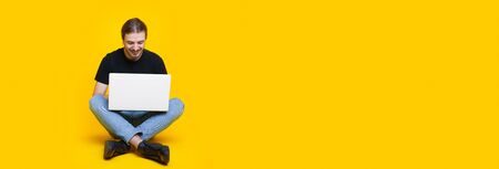 Bearded caucasian man with long hair is sitting on a yellow wall with free space and working at the...
