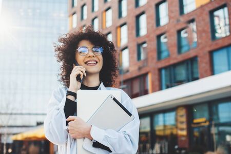 Lovely caucasian businessperson with curly hair is talking on phone while posing with some modern...