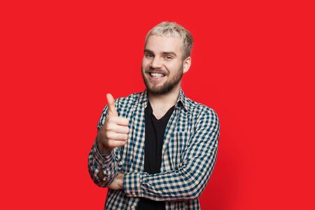 Bearded caucasian man with blonde hair is gesturing the like sign on a red background