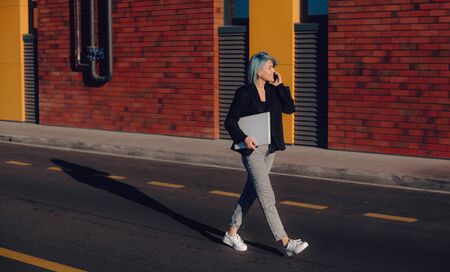 Blue haired caucasian student walking in the street while having a phone discussion and holding a laptopの写真素材