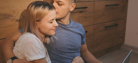 Close up portrait of a caucasian blonde woman kissed by her lover while sitting on the floor in the kitchen and using a laptopの写真素材