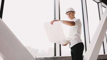 Young engineer holding a building plan and posing upstairs with a helmet