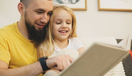 Cheerful blonde girl and her father with beard reading together a bookの写真素材