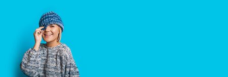 Nice woman with blue hat and blonde hair touching it while smiling at camera on a blue wall with...
