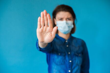 Charming caucasian woman with blue jeans shirt wearing a medical mask gesturing stop sign on a studio wallの写真素材