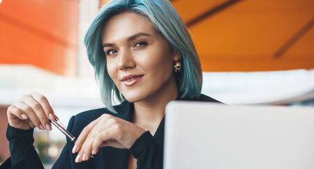 Lovely middle aged woman with blue hair working in a cafeteria with a laptop while using a pen and looking at cameraの写真素材