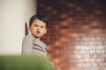 Sad caucasian boy with black hair sitting up on the sofa with a stone wall on backgroundの写真素材