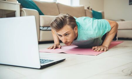 Caucasian man doing pushups on a sport carpet while looking at the laptop on the floor