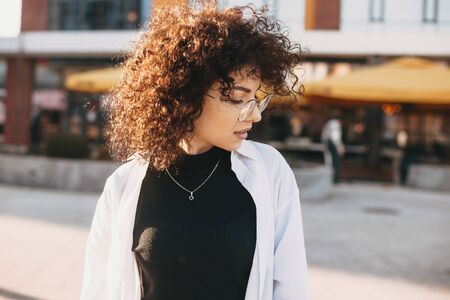 Close up photo of a young woman with curly hair posing outside in a sunny day while looking downの写真素材