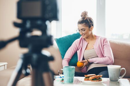 Young woman vlogging with a cup of tea and sandwiches on table while wearing eyeglasses and holding...