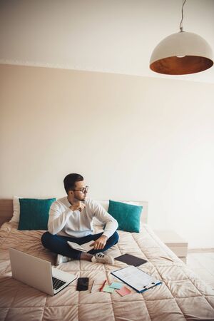 Thinking caucasian man with beard and eyeglasses is working in bed with a computer and documentsの写真素材