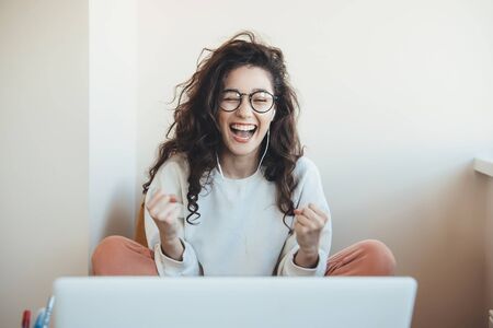 Lovely young woman with eyeglasses and curly hair smiling and gesturing the win emotions in front of the laptop at homeの写真素材