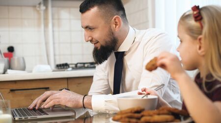 Caucasian businessman with beard working at the computer and his daughter eating cookies at the same tableの写真素材