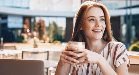 Freckled caucasian girl drinking a milkshake is working at the laptop in a cafeteriaの写真素材