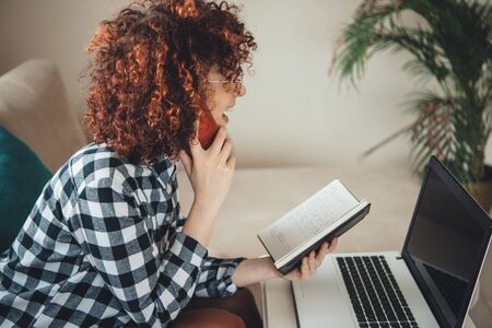 Happy caucasian lady with curly hair talking on mobile and using a laptop while looking in the copybookの写真素材