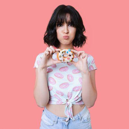 Charming brunette lady in fashionable clothes holding a tasty colorful donut and posing on a pink studio wallの写真素材