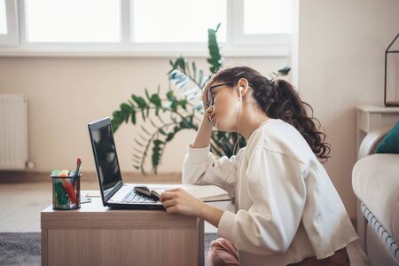 Curly haired caucasian woman tired of lessons wearing eyeglasses and headphones doing homework at...