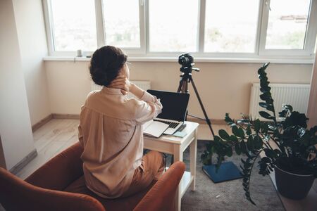 Tired caucasian woman in formal wear having online meeting using a camera and laptop while sitting at the table in armchairの写真素材