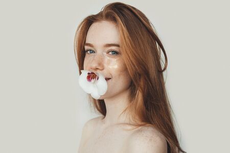 Charming ginger lady with freckles   shoulders holding a flower with her mouth and posing on a white...