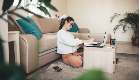 Side view photo of a concentrated caucasian woman with curly hair and eyeglasses writing something using a laptopの写真素材